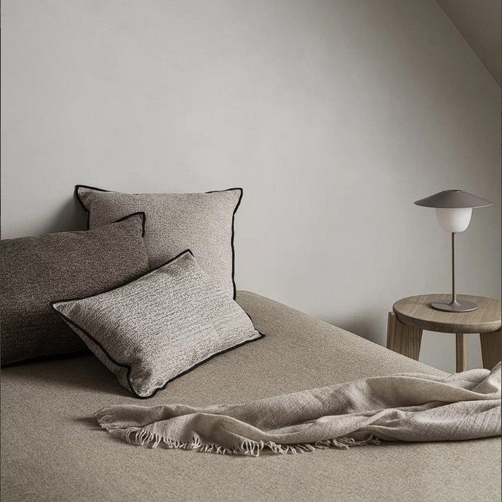 A bed stacked with square shaped velvet cushions next to a nightstand.