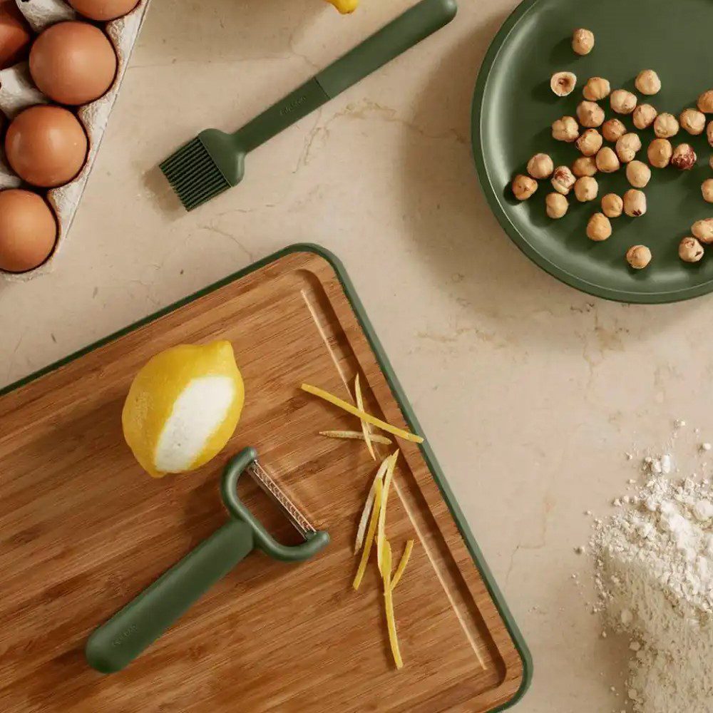 Top view of a kitchen counter with a julienne peeler producing thinly-sliced lemon rinds placed on a chopping board and with other utensils on the side.