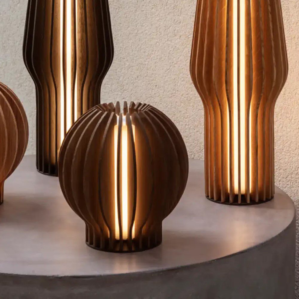 Natural light, perspective view of four bulb- and canister-shaped lit wooden oak lamps placed on top of a table.