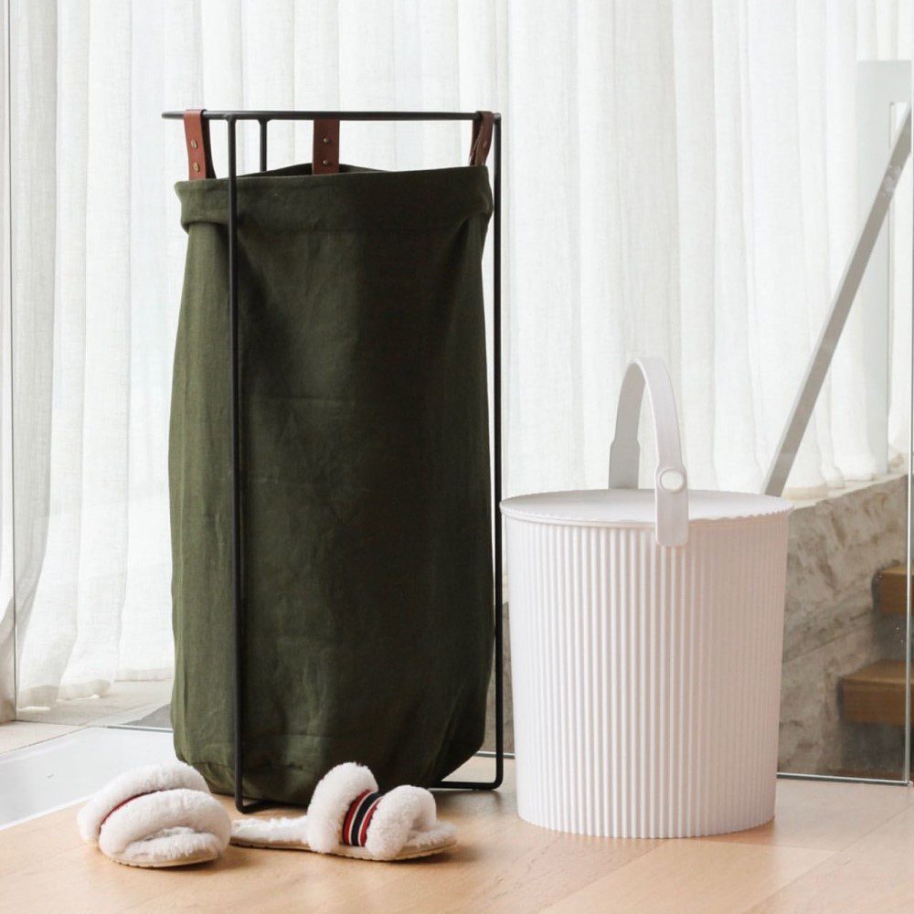 Natural light, perspective photo of a tall black laundry hamper, a white storage bin with its handle upright, and a pair of faux fur slippers beside the laundry hamper.