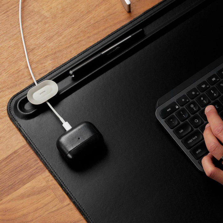 Natural light, top view of a headphone charging case placed on the top left corner of a black, rectangular leather desk mat.