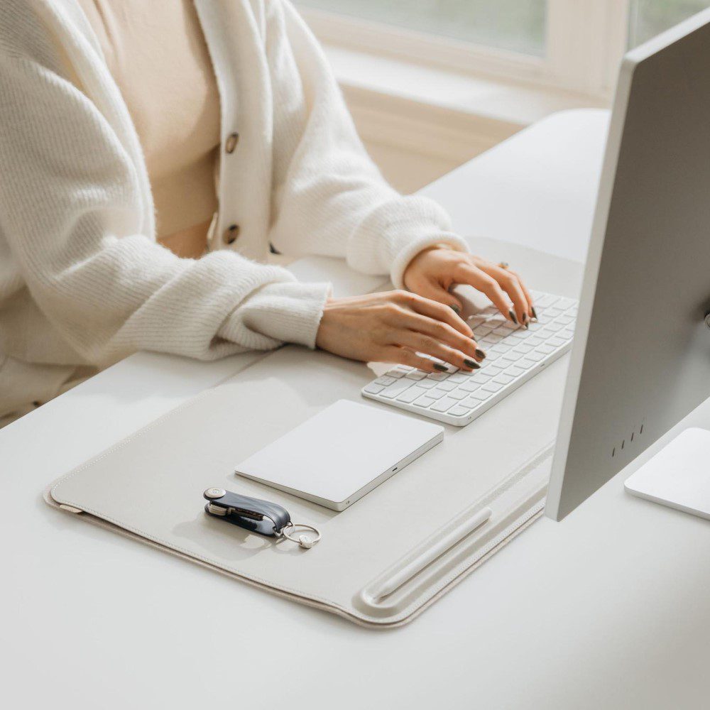 Studio lighting, perspective view of person typing on a keyboard placed on top of a rectangular-shaped, light grey desk mat facing a computer screen.