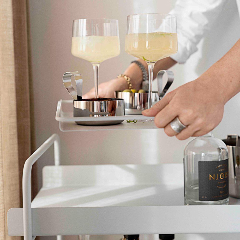 Sun-lit, cropped view of a person taking and holding two cocktail drinks and serving bowls on a tray away from the top of a cocktail trolley.