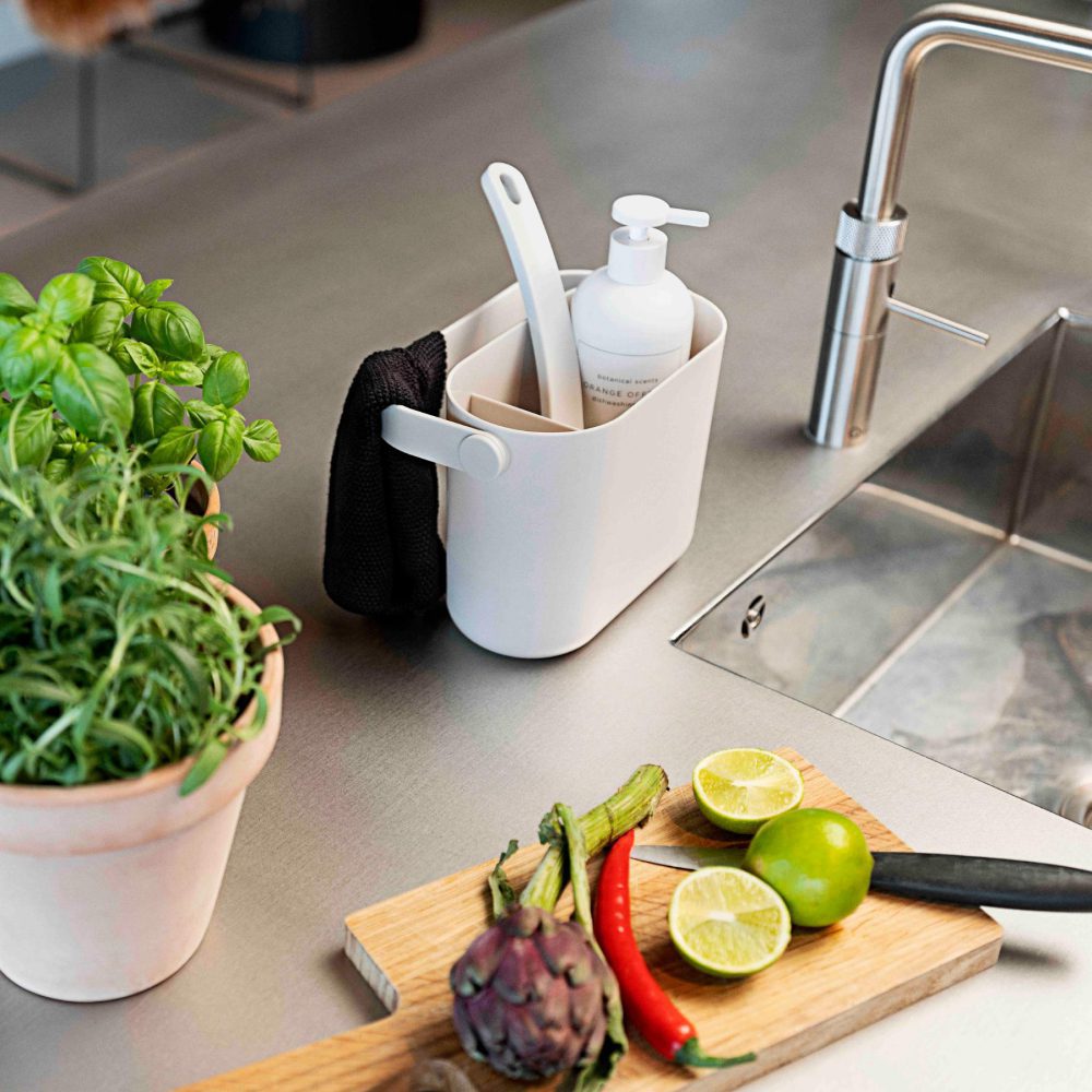 Natural light, isometric view of a stainless steel kitchen sink with a grey-coloured tray of cleaning tools beside the sink's faucet and a chopping board with sliced fruits and vegetables.